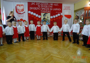 Na tle biało czerwonej dekoracji stoją wpółkolu dzieci ubrane na biało-czerwono.